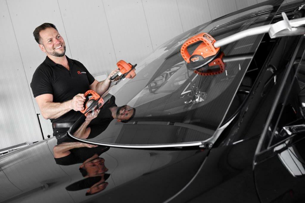 Ein Mechaniker der Autowerkstatt Ackermann bearbeitet die Windschutzscheibe eines schwarzen Autos mit speziellen Werkzeugen für Autoglas-Dienstleistungen in der Werkstatt.