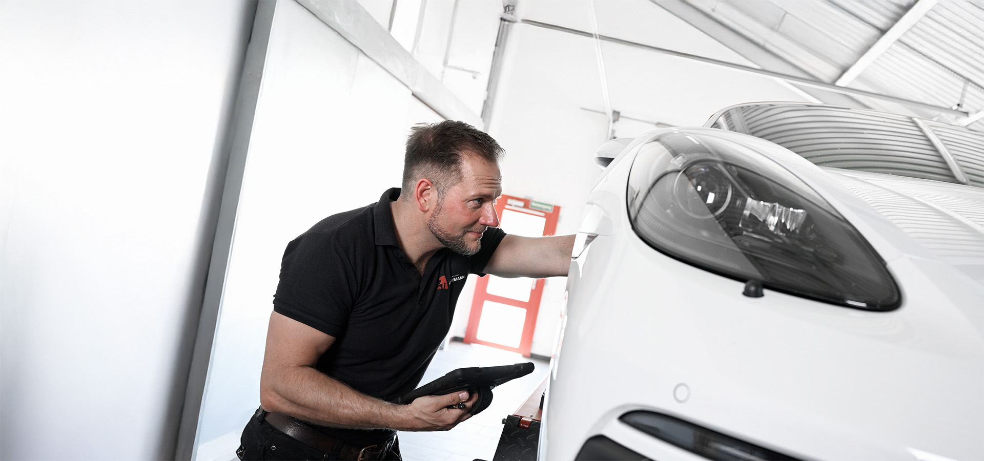 Ein Mechaniker der Autowerkstatt Ackermann prüft in der Werkstatt ein weißes Auto mit einem Tablet – das Fachbetrieb bietet Werkstattservice, Lackiererei, Unfallinstandsetzung und Leistungen für Young- und Oldtimer.