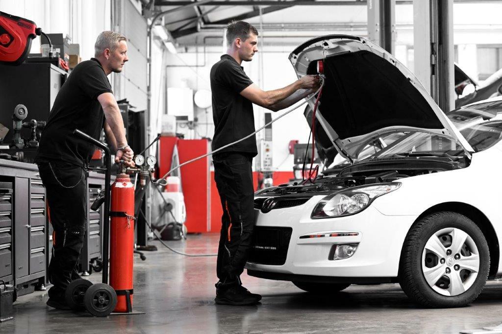 Zwei Mechaniker führen in einer KFZ-Werkstatt Reparaturarbeiten an einem weißen Auto mit geöffneter Motorhaube durch, wobei einer das Motorwerkzeug bedient und der andere eine rote Ausrüstung nutzt.