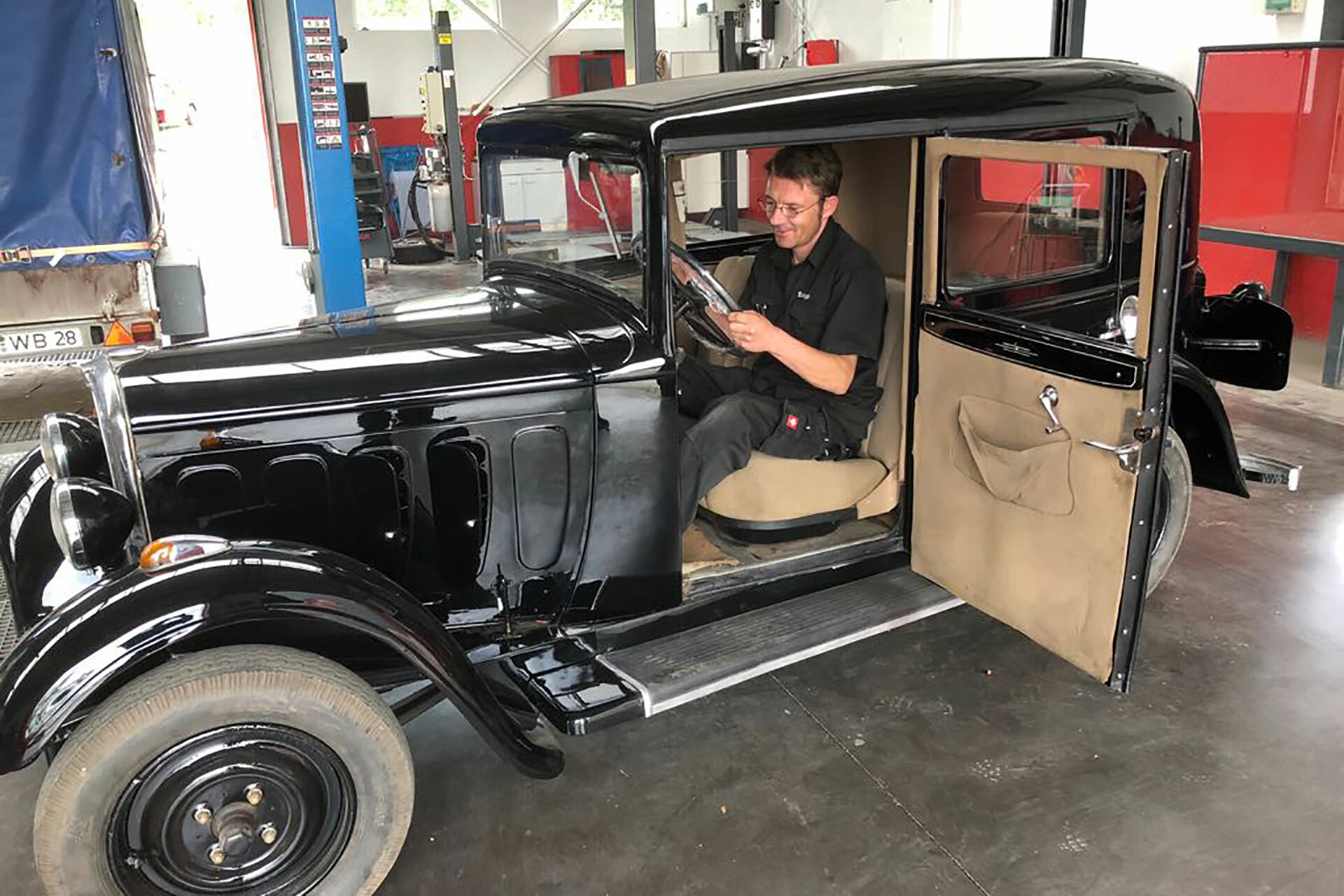 Ein Mechaniker sitzt in einem schwarzen Oldtimer mit geöffneter Tür in der Werkstatt von Autowerkstatt Ackermann, einem KFZ-Fachbetrieb, der Werkstattservice und Leistungen für Young- und Oldtimer unter einem Dach vereint.