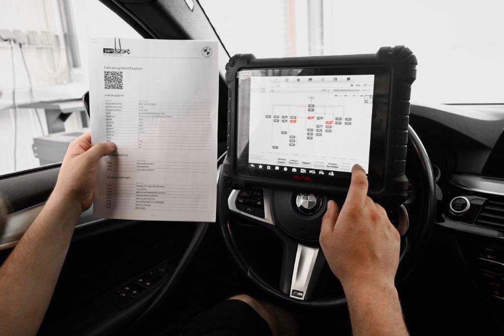 Ein Mitarbeiter der Autowerkstatt Ackermann nutzt ein Diagnosegerät, um die Fahrzeugdaten eines BMW auszulesen, und hält dabei ein Dokument mit Fahrzeugidentifikation in der Hand.