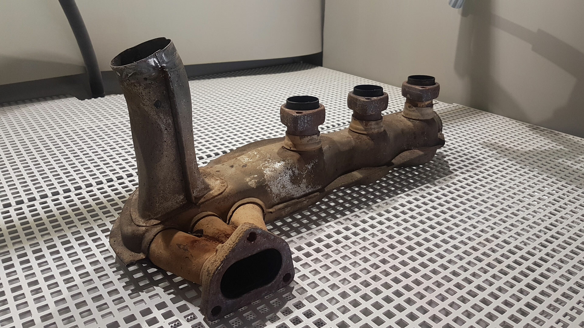 Verrosteter Auspuffkrümmer mit mehreren Anschlüssen – ein KFZ-Teil mit Korrosionserscheinungen, das Reparaturen oder Instandsetzungen in einer Autowerkstatt erfordert.