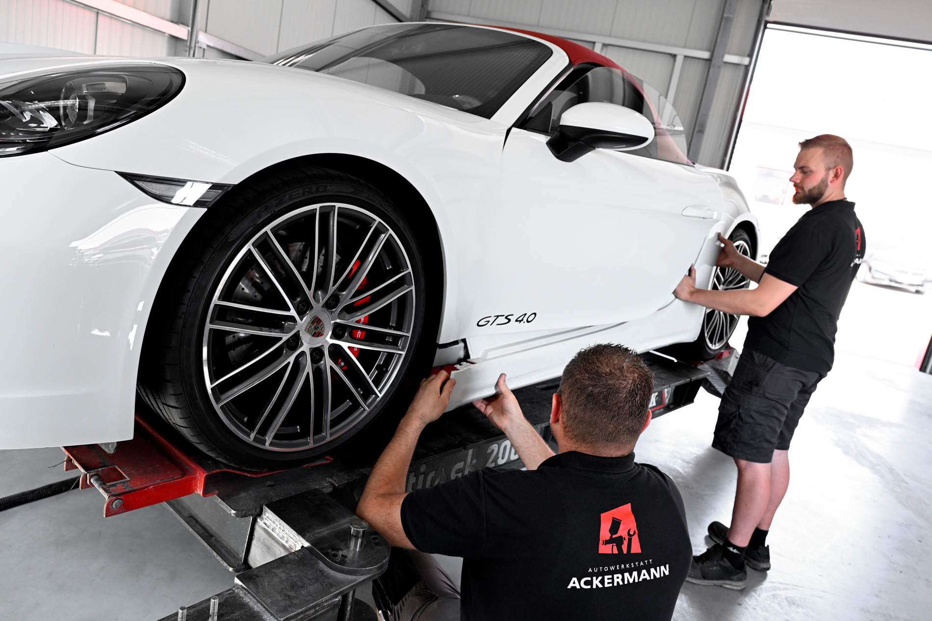 Zwei Mechaniker von Autowerkstatt Ackermann arbeiten an einem weißen Porsche 718 GTS 4.0 auf einer Fahrzeughebebühne in der Werkstatt.