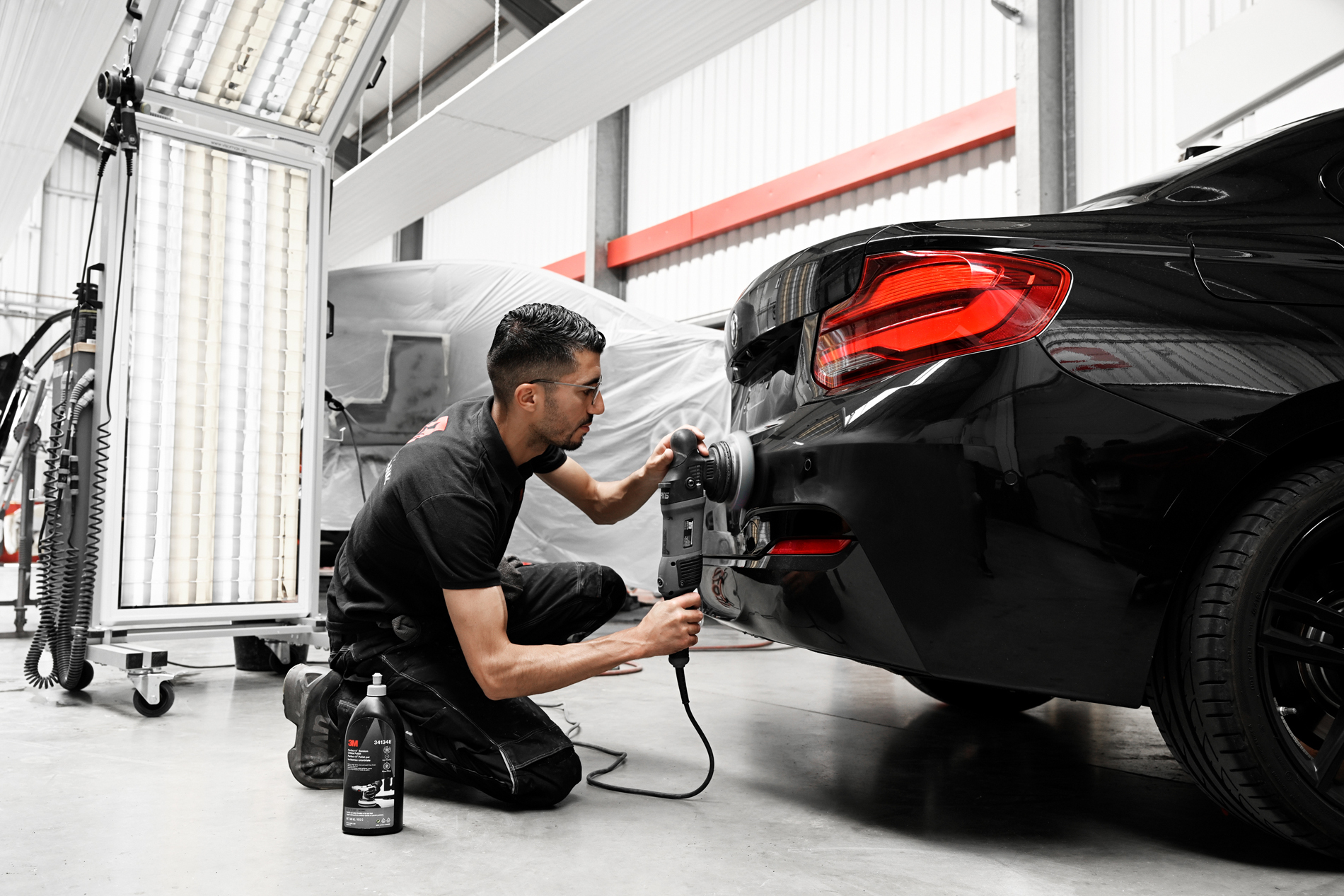 Mechaniker bei Autowerkstatt Ackermann poliert das Heck eines schwarzen Autos mit einem Poliergerät, wobei ein 3M-Produkt zum Einsatz kommt, in einer professionellen Werkstattumgebung.