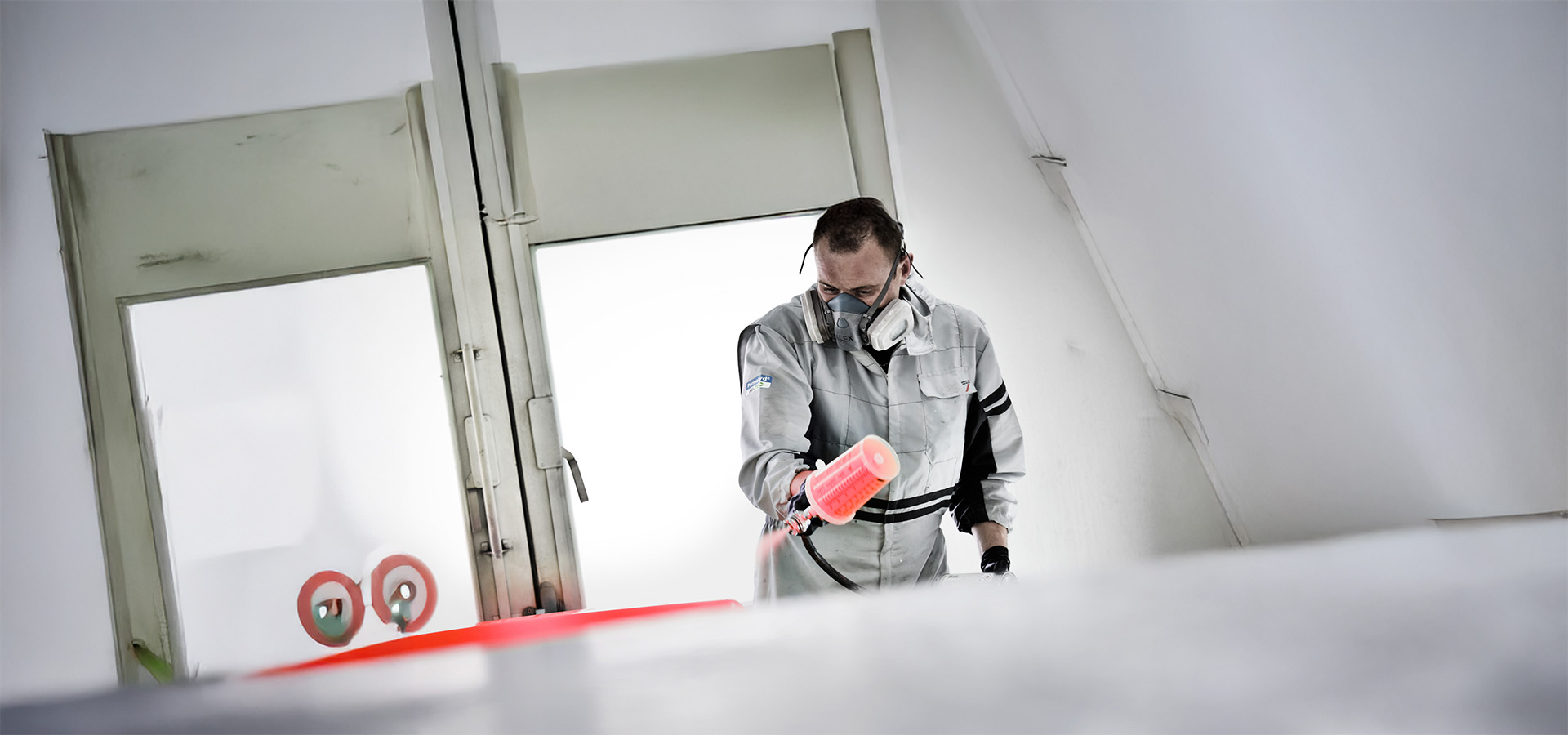 Ein Mitarbeiter der Autowerkstatt Ackermann lackiert ein Fahrzeugteil mit einem Spritzgerät in der Werkstatt, dabei trägt er Schutzkleidung und Atemschutz für eine fachgerechte und saubere Ausführung.