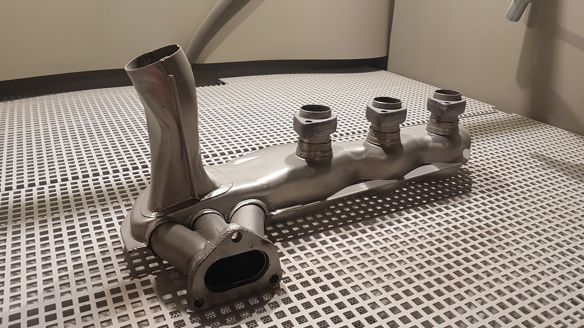Sandgestrahltes Getriebe-Teil mit mehreren Anschlüssen, metallisch und auf einem Gitterboden platziert, typisch für die fachgerechte Fahrzeuginstandsetzung in einer KFZ-Fachwerkstatt.