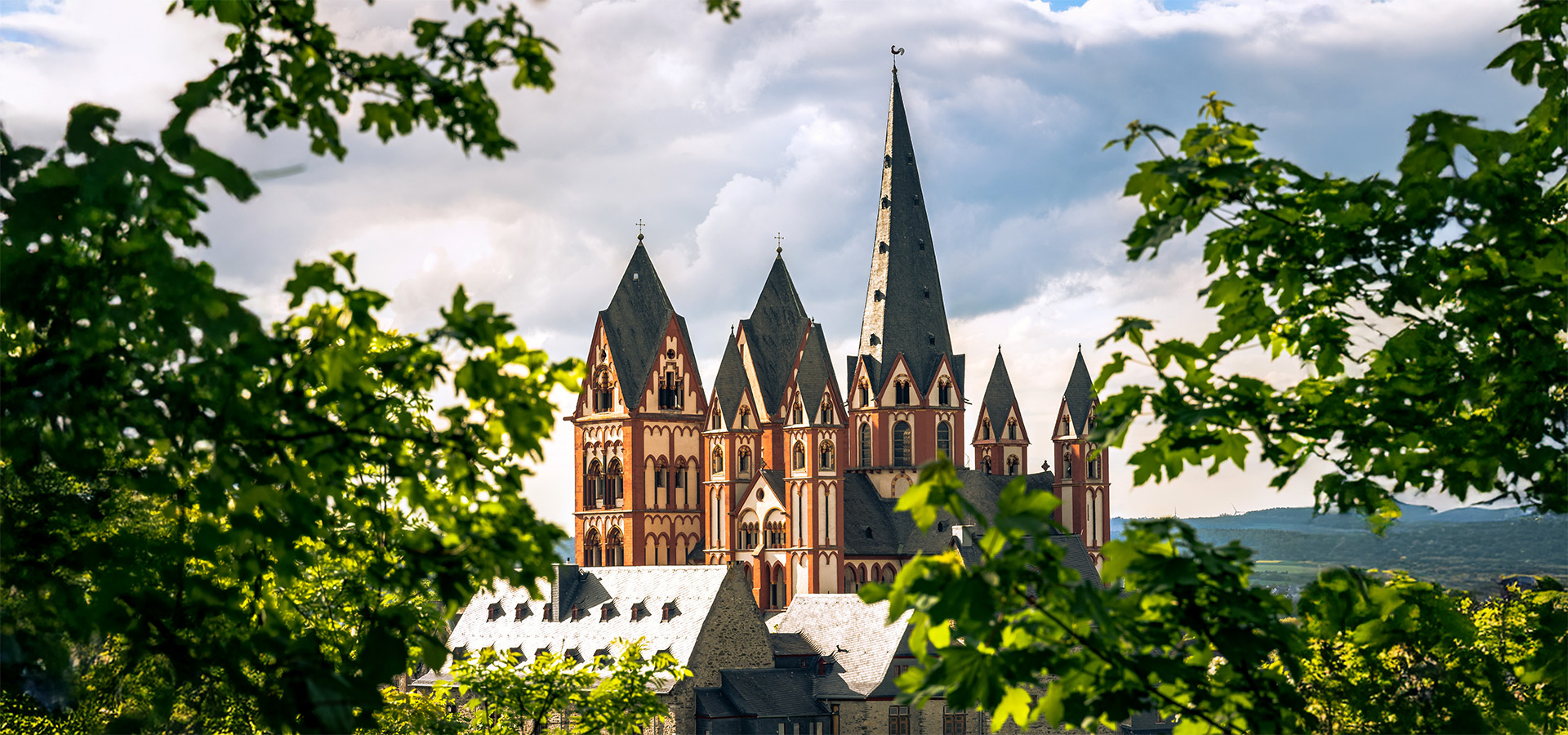 Der Limburger Dom mit seinen markanten gotischen Spitztürmen ragt hinter frischem Laub gegen einen wolkenreichen Himmel.