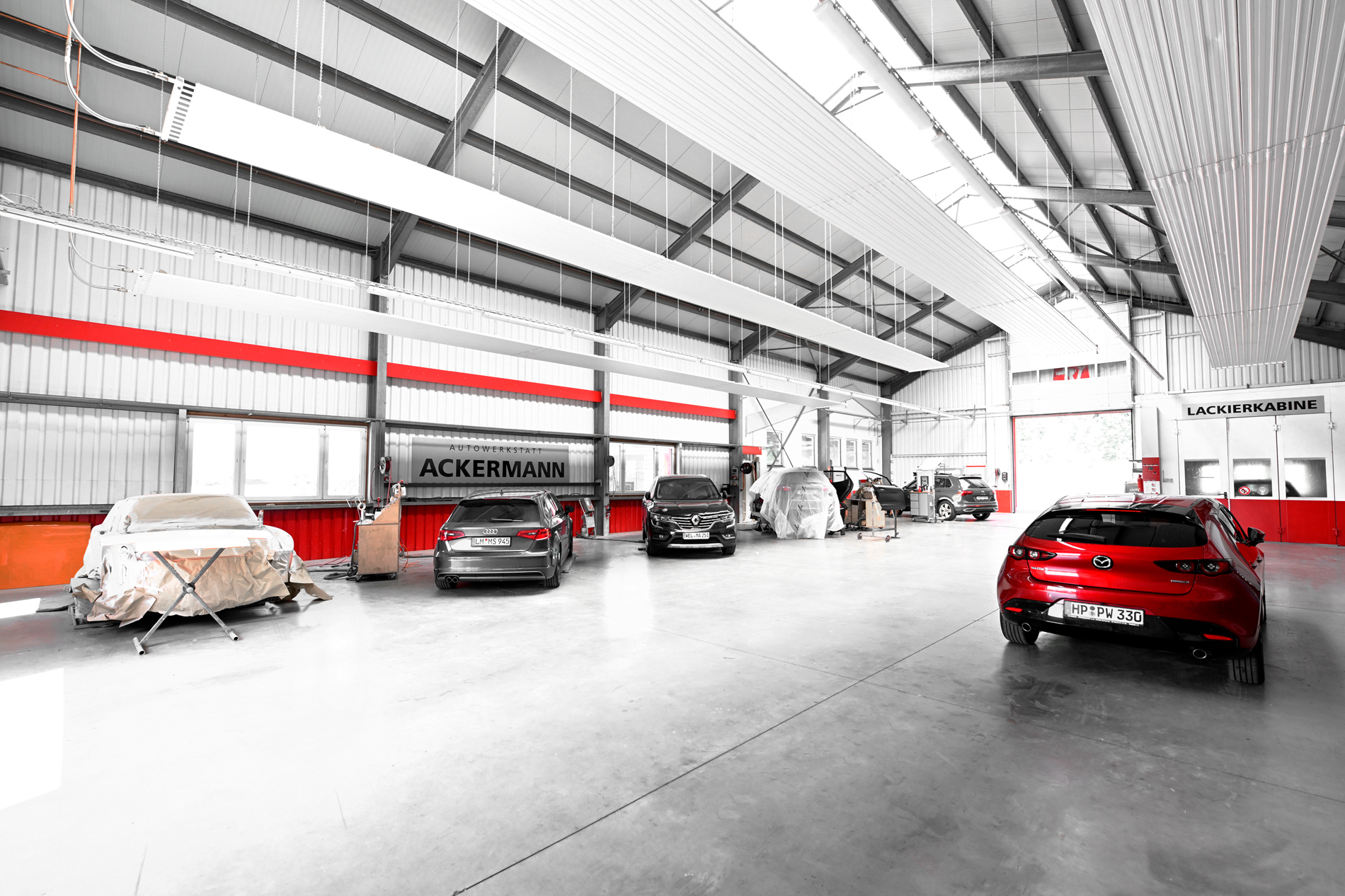 Innenraum der Autowerkstatt Ackermann mit mehreren Fahrzeugen, darunter einem roten Mazda, abgedeckten Autos und der Lackierkabine im Hintergrund, die Werkstattservice, Lackierung und Fahrzeugreparaturen unter einem Dach anbietet.