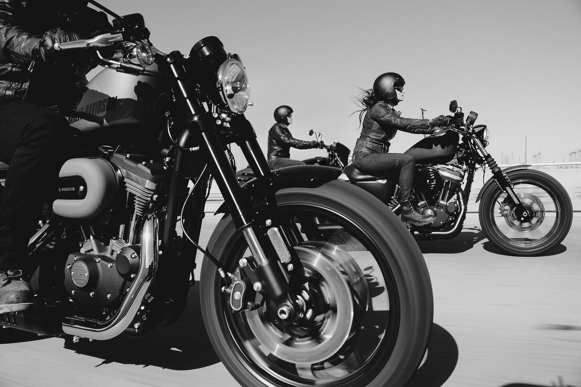 Drei Motorradfahrer fahren auf einer Straße, darunter ein Harley-Davidson im Vordergrund, in Schwarz-Weiß-Fotografie.